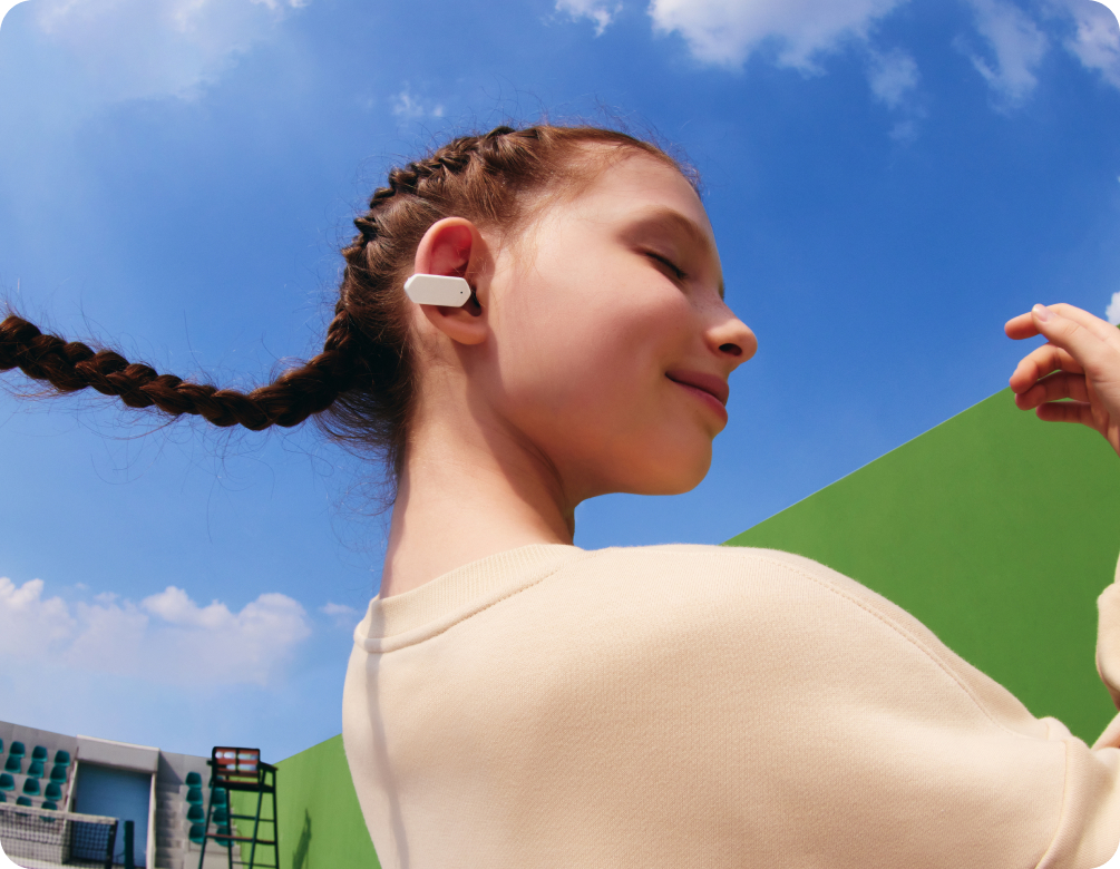 Mädchen mit Zöpfen und weißem imoo Openbuds E5 im Ohr lächelt im Freien vor blauem Himmel